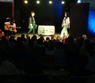 Delar av publiken syns i förgrunden av en scen där man spelar teater